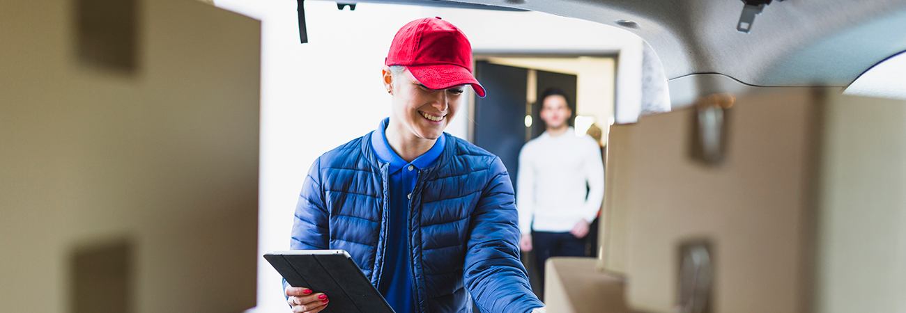 A smiling delivery worker in a red cap and blue jacket checks moving boxes with a tablet while a customer watches.