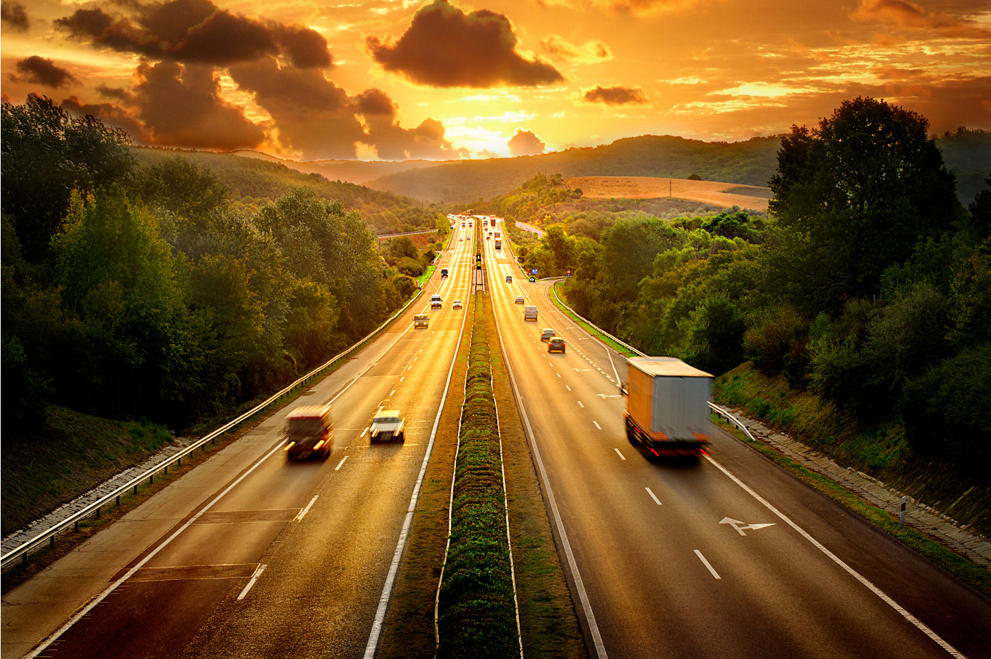 Busy highway at sunset with cars and trucks driving through a scenic, tree-lined landscape.