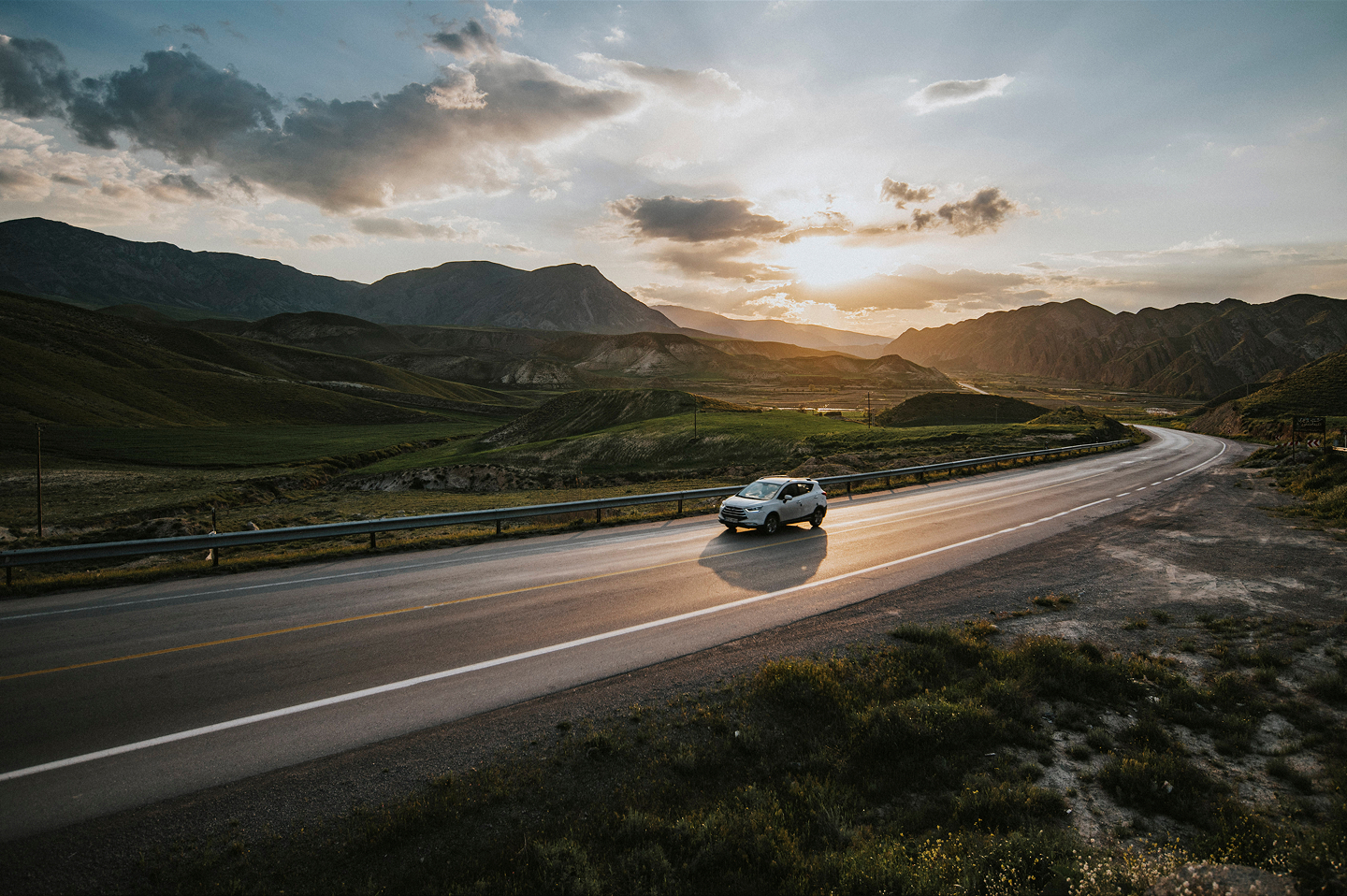 Car driving on a winding highway through rolling green hills with a sunset over distant mountains.