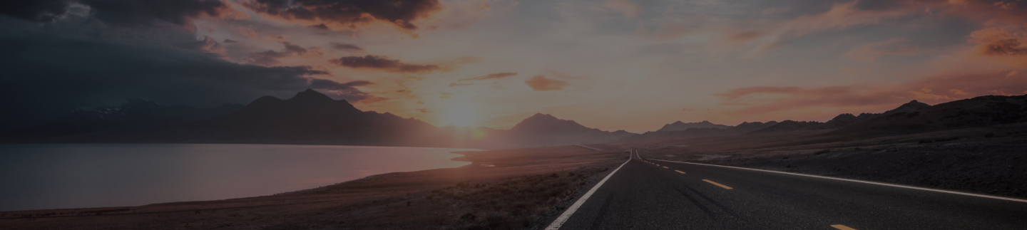 Scenic open road alongside a lake with mountains in the background at sunset.