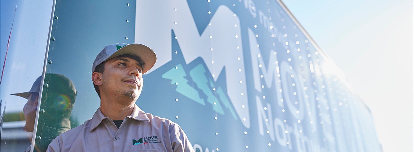 Move Northwest employee in a uniform and cap standing beside a branded Moving Truck with a reflective surface. Moving in somewhere in Washington State local or interstate.